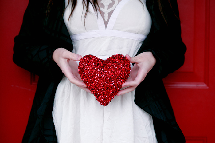 Female holding red glitter heart wearing white shirt and black jacket in front of red door