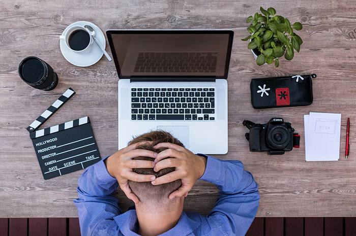 Man with hands on head leaning over desk in front of laptop with cup of coffee camera and movie action sign either side of him