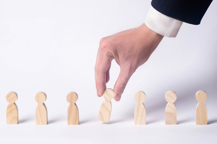 Man's hand in business suit attire selecting wooden cutout of person's silhouette in a line of 7