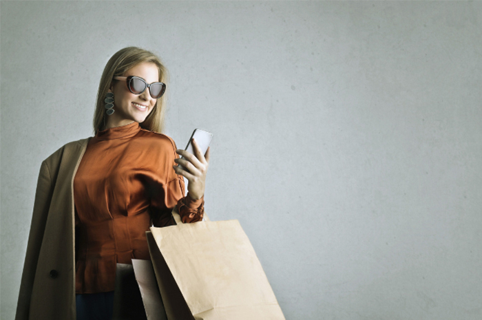 Female shopper wearing beautiful clothing and sunglasses with big smile looking at her phone holding a brown paper shopping bag