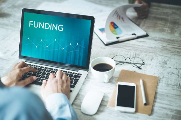 Business man typing words FUNDING into laptop computer on desk with black coffee phone glasses pen folder and charts nearby