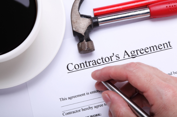 Contractor's agreement on desk with person holding pen near cup of coffee and other tools including hammer