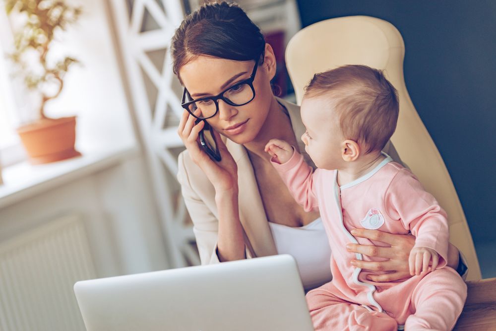 Mother working on laptop speaking on phone holding little baby