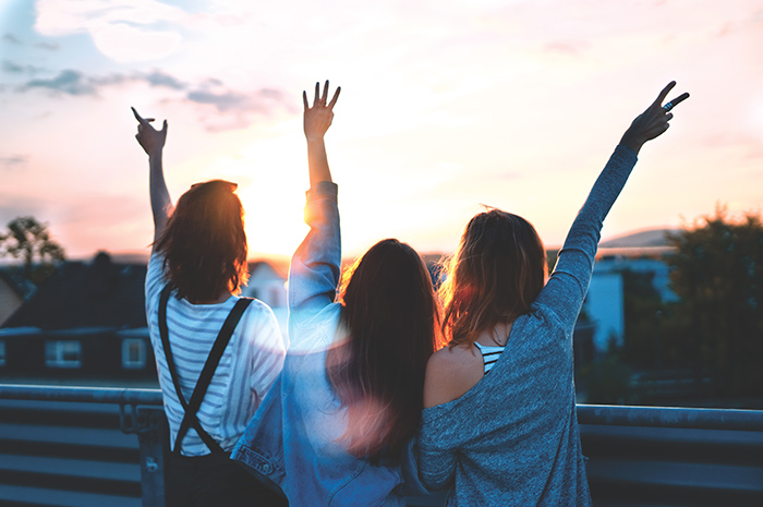 Three women facing sunrise on balcony with hands in the air