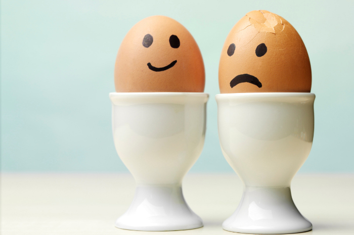 Two eggs in egg cups one with a happy face and the other with a sad face and slightly cracked shell