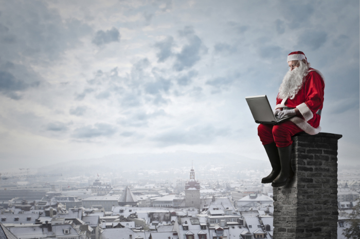 Santa Claus sitting on chimney working on laptop computer above skyline