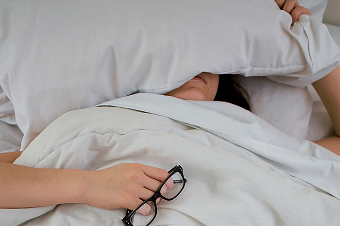 Woman in bed covering half of face with pillow holding black glasses in hand
