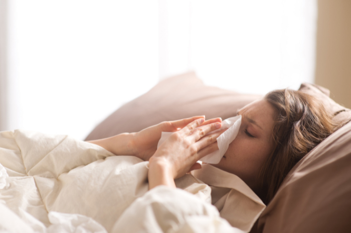 Woman laying down in bed blowing her nose with a tissue looking very unwell and sick
