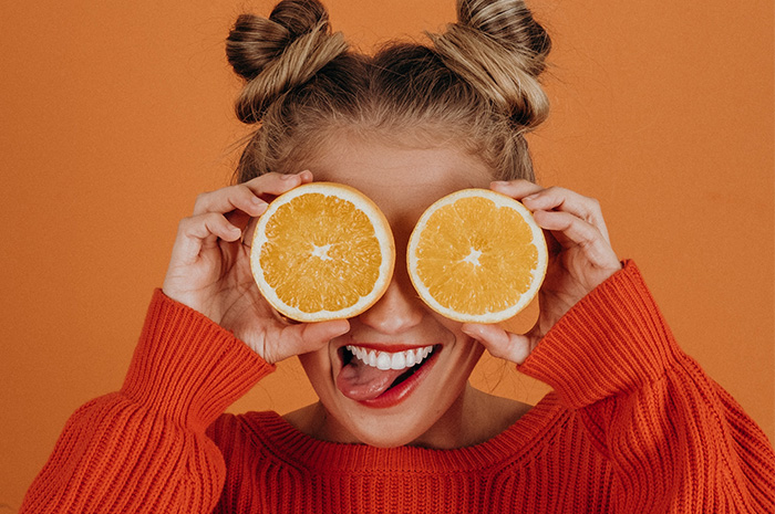 Female in orange jumper holding half an orange over each eye hair in two little buns